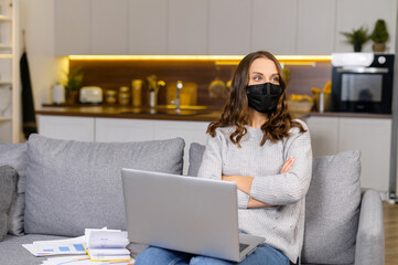 Self isolation concept. A young woman wearing protective face mask sits on the couch with a laptop,...