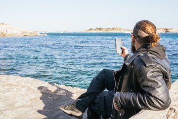Caucasian man with beard, long hair and sunglasses sitting by the sea at sunset and taking a selfie with his smartphone in a cove in Portals. Palma de Mallorca, Spain (Copyspace)