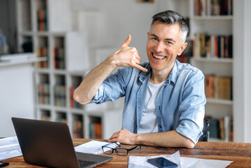 Friendly sociable confident middle aged gray-haired Caucasian male freelancer, in stylish clothes, works remotely at a laptop, shows the call me gesture, looks at the camera, smiles