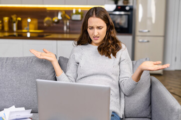 Confused female entrepreneur sits on the couch and staring at the laptop screen, young woman shrugs...