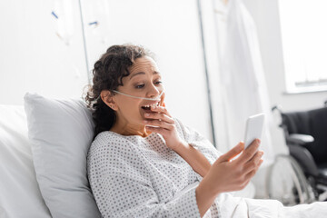 astonished african american woman covering mouth while looking at cellphone in clinic