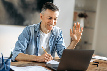 Cheerful handsome successful mature Caucasian businessman, freelancer or manager in stylish clothes, using laptop, talking to colleagues or clients on a video call, greeting with hand gesture, smiles