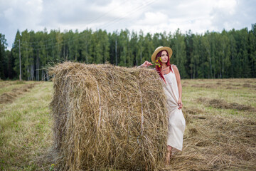 Naklejka na ściany i meble A girl with red long hair stands by a haystack in a field