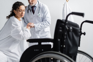 doctor supporting african american woman near wheelchair on blurred foreground