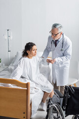 Obraz premium doctor helping african american woman getting up from bed near wheelchair in hospital
