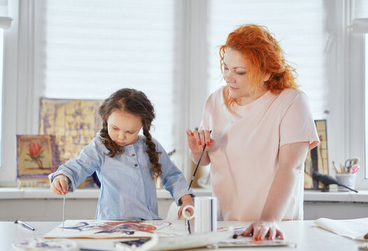 Family leisure activity at home concept. Mother and small daughter drawing together using paintbrushes and watercolors or acrylic. Young woman helping or teaching daughter sitting on a table to paint