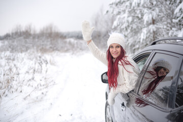 Fototapeta na wymiar A woman in a white hat and sweater in the winter from the car waves her hand in a white mitten and wishes a happy journey. winter christmas trip by car. Merry christmas and new year