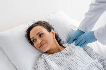 doctor in latex gloves touching shoulder of smiling african american woman lying in hospital bed