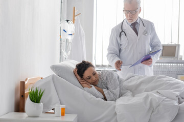 Fototapeta premium middle aged doctor with clipboard standing near upset african american woman lying in hospital bed