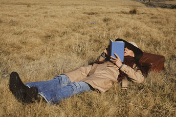 Woman traveler with hat and suitcase is lying down in a golden grass field and is reading a book.