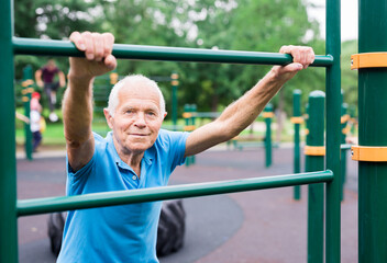 mature cheerful pensioner man doing physical exercises on sports equipped playground