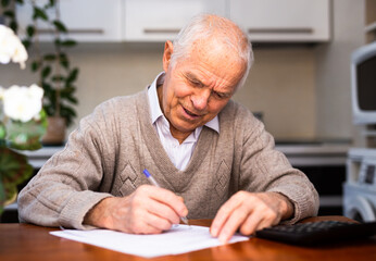 elderly pensioner writing on piece of paper at table in kitchen - obrazy, fototapety, plakaty