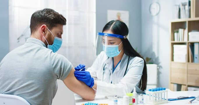 Close up of young Asian beautiful female doctor in medical mask and face shield doing injection with covid-19 vaccine in hospital to handsome Caucasian male patient. Vaccination, coronavirus cure