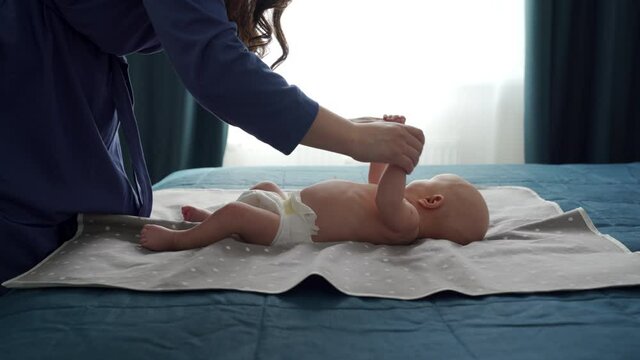Mother with baby boy doing exercise simple exercises to make baby bones and muscles stronger, newborn infant lying on bed against the window. High quality 4k footage