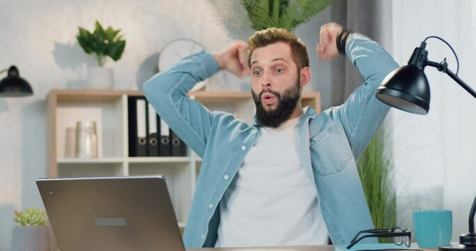 Handsome happy satisfied emotional young bearded guy recieving good news on computer and enjoying from results with raised hands
