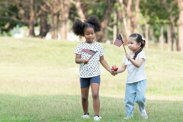 African and Caucasian kids playing together and holding small America flag and red apple at green park. Ethnic diversity of relationship with little girls at 4th of July concept