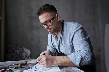 Bearded attractive brunette man in apron and eyeglasses making linocut sketch using felt pen....