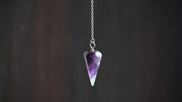 Purple Amethyst crystal pendulum hanging on chain on dark background.