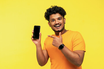 Handsome and confident Indian smiling guy isolated on yellow background with modern phone with black screen in hand, looking at camera