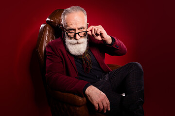 Portrait of serious person sit in leather chair arm touch glasses look camera isolated on dark red...