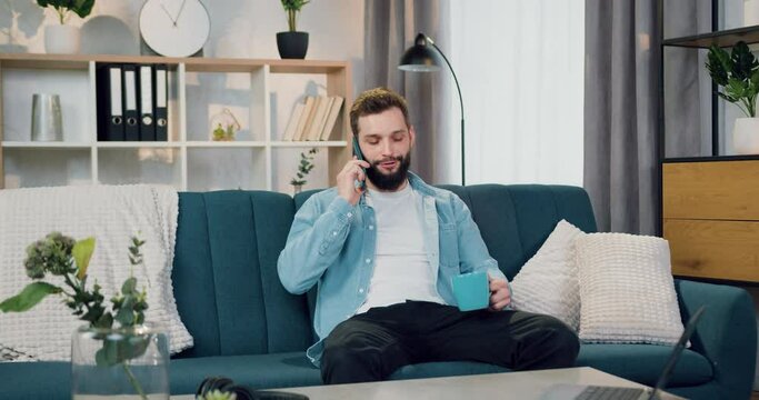 Portrait of handsome in good mood calm young berded guy which sitting on the couch at home and talking on smartphone simultaneously drinking coffee