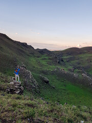 A man standing on a green hill in Xilinhot, Inner Mongolia and enjoys a sunset. He is wearing a cowboy hat. Endless grassland. The sun starts setting behind the horizon, coloring the sky orange