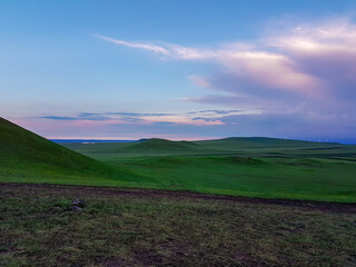 A panoramic view on a hilly landscape of Xilinhot in Inner Mongolia. Endless grassland with a few wind turbines in the back. The sun starts to set, coloring the sky pink. Thick, rainy clouds.
