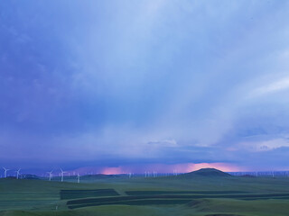 A panoramic view on a hilly landscape of Xilinhot in Inner Mongolia. Endless grassland with a few wind turbines in the back. The sun starts to set, coloring the sky pink. Thick, rainy clouds.