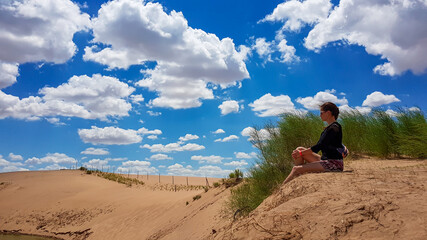 Naklejka na ściany i meble A woman in shorts sitting on a sand dune on Hunshandake Desert in the nearby of Xilinhot, Inner Mongolia. The sand dunes are overgrown with a bit of grass. Blue sky with thick, white clouds. Solitude