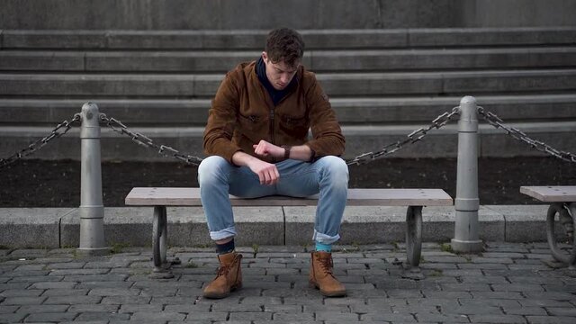 Young man sitting on a city bench,checking his watch,waiting,