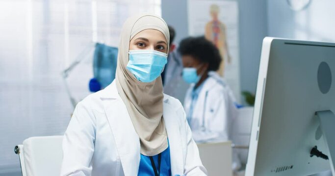 Close up portrait of beautiful Arabic young female professional physician sitting at workplace at desk in cabinet looking at camera. Work during covid-19 pandemic. Hospital, clinic concept