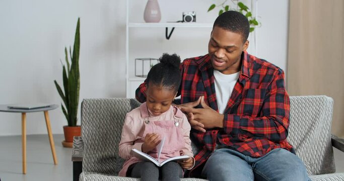 Afro american father sits with beloved daughter on couch in interior of modern living room kid holds book album in gray cover turns pages looks pictures in literature with interest, child development