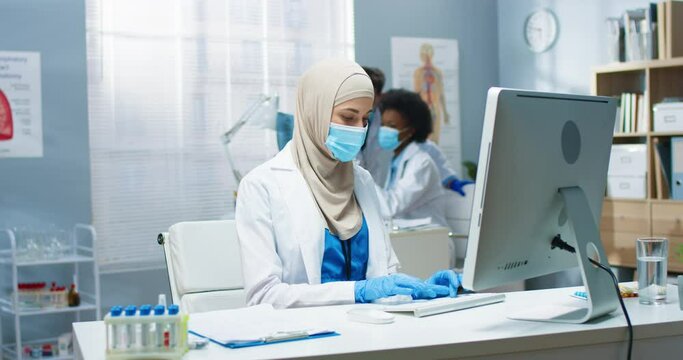 Young Arab beautiful professional female physician in medical mask working, typing and browsing online on computer sitting at workplace. Covid-19 virus, healthcare, medicine concept