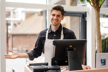 Smiling young man in apron standing at the cash register - obrazy, fototapety, plakaty
