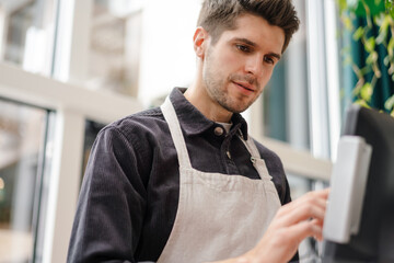 Serious young man in apron standing at the cash register