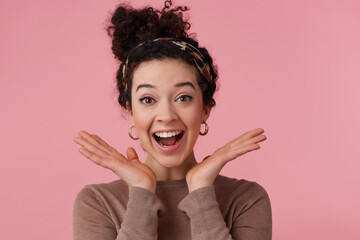 Portrait of attractive, joyful girl with dark curly hair bun. Wearing headband, earrings and brown sweater. Has make up. Keeps palms spread. Watching at the camera isolated over pastel pink background
