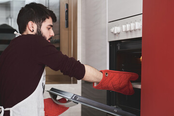 Young caucasian man in apron cooking in a kitchen. He is removing the food from the oven.