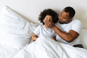 Fototapeta premium Black father covering his son's mouth while lying in bed at home