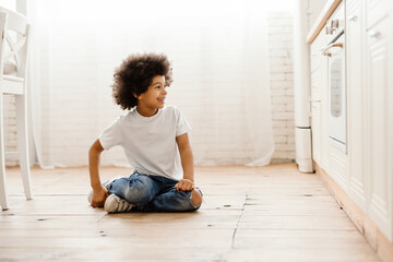Black curly boy smiling while sitting on floor in home kitchen