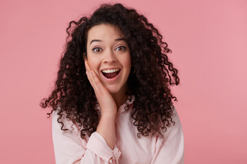 Amazed woman, beautiful girl with dark curly hair bun. Wearing earrings and pastel pink shirt. Has make up. Touching her face in surprise. Watching at the camera isolated over pastel pink background