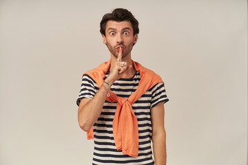 Portrait of shocked male with brunette hair and bristle. Wearing striped t-shirt and orange sweater tied on shoulders. Shows silence sign. Watching at the camera isolated over grey background