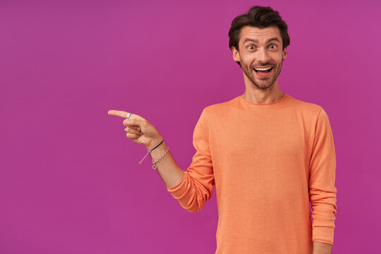 Surprised man with brunette hair and bristle. Wearing orange sweater. Has bracelets and rings. Watching at the camera and pointing finger to the left at copy space, isolated over purple background