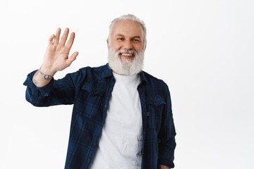 Friendly old man waving hand to say hello. Smiling senior guy with tattoos waves as greeting, making hi gesture, standing happy against white background