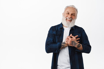 Thank you. Happy senior tattooed man, holding hands on heart and smiling, being flattered or touched, looking heartfelt, pleased by something, standing over white background