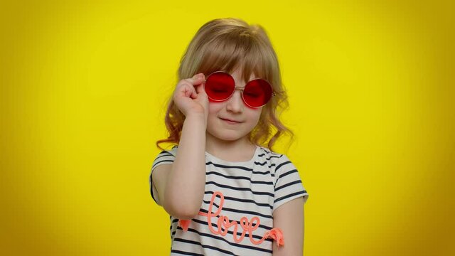 Happy playful little blonde teen kid child girl in sunglasses blinking eye, looking at camera with toothy smile, winking and flirting, expressing optimism. Young children on yellow studio background