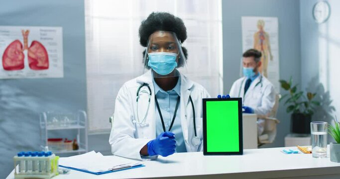 Portrait of African American female physician sitting in hospital in protective face shield in covid pandemic holding in hand tablet with chroma key speaking on video consultation looking at camera