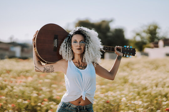 Chica delgada con pelo muy rizado y tatuajes posando con guitarra acústica en un campo de margaritas