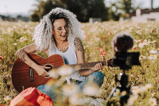 Chica guapa delgada con el pelo muy rizado tocando la guitarra en un campo de margaritas mientras se graba un vlog