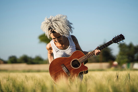Chica guapa tatuada con pelo muy rizado posando en un campo de trigo con una guitarra acsutica