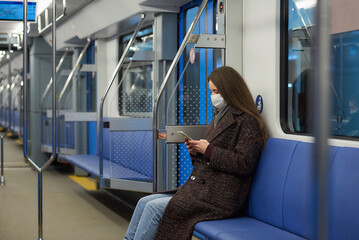 A woman in a face mask is sitting and using a smartphone in a modern subway car.